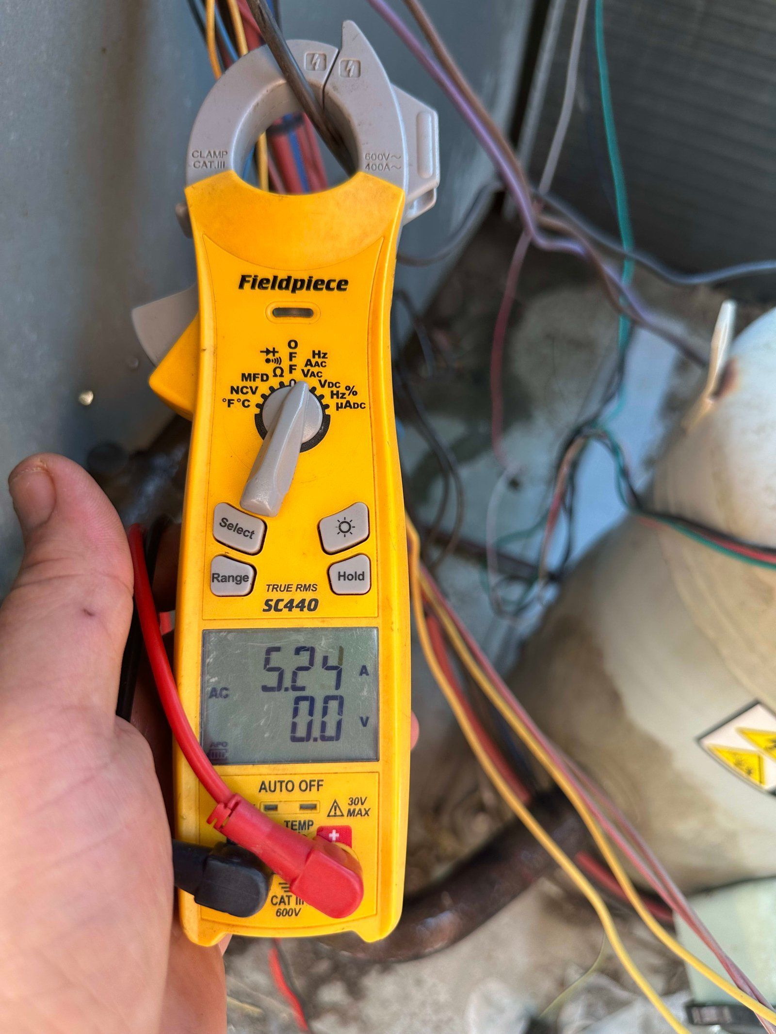 A person using a yellow Fieldpiece multimeter to measure electrical current on HVAC wiring; the display reads 5.24.