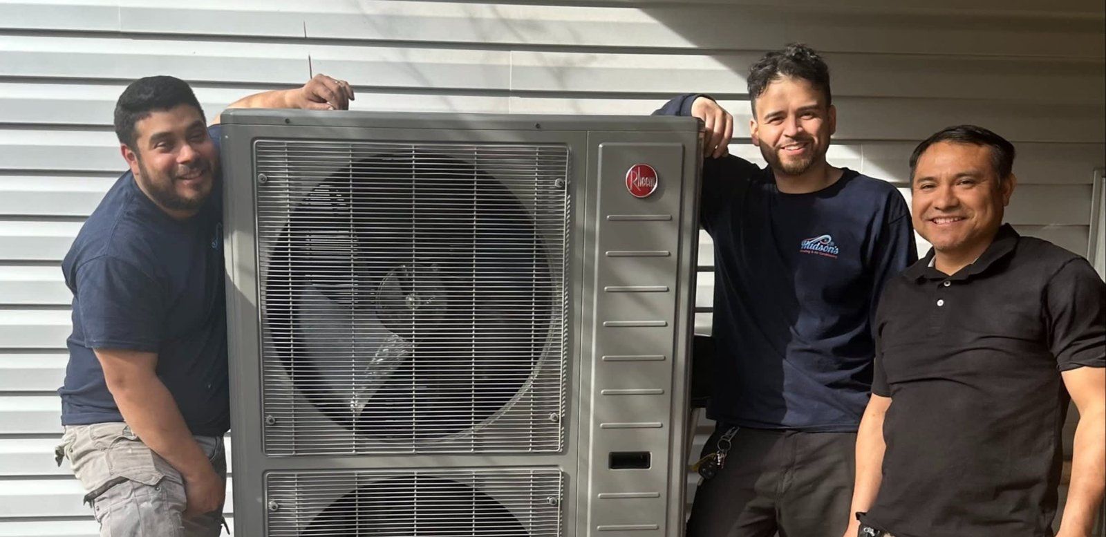 Three men pose with a large air conditioning unit in front of a gray wall.