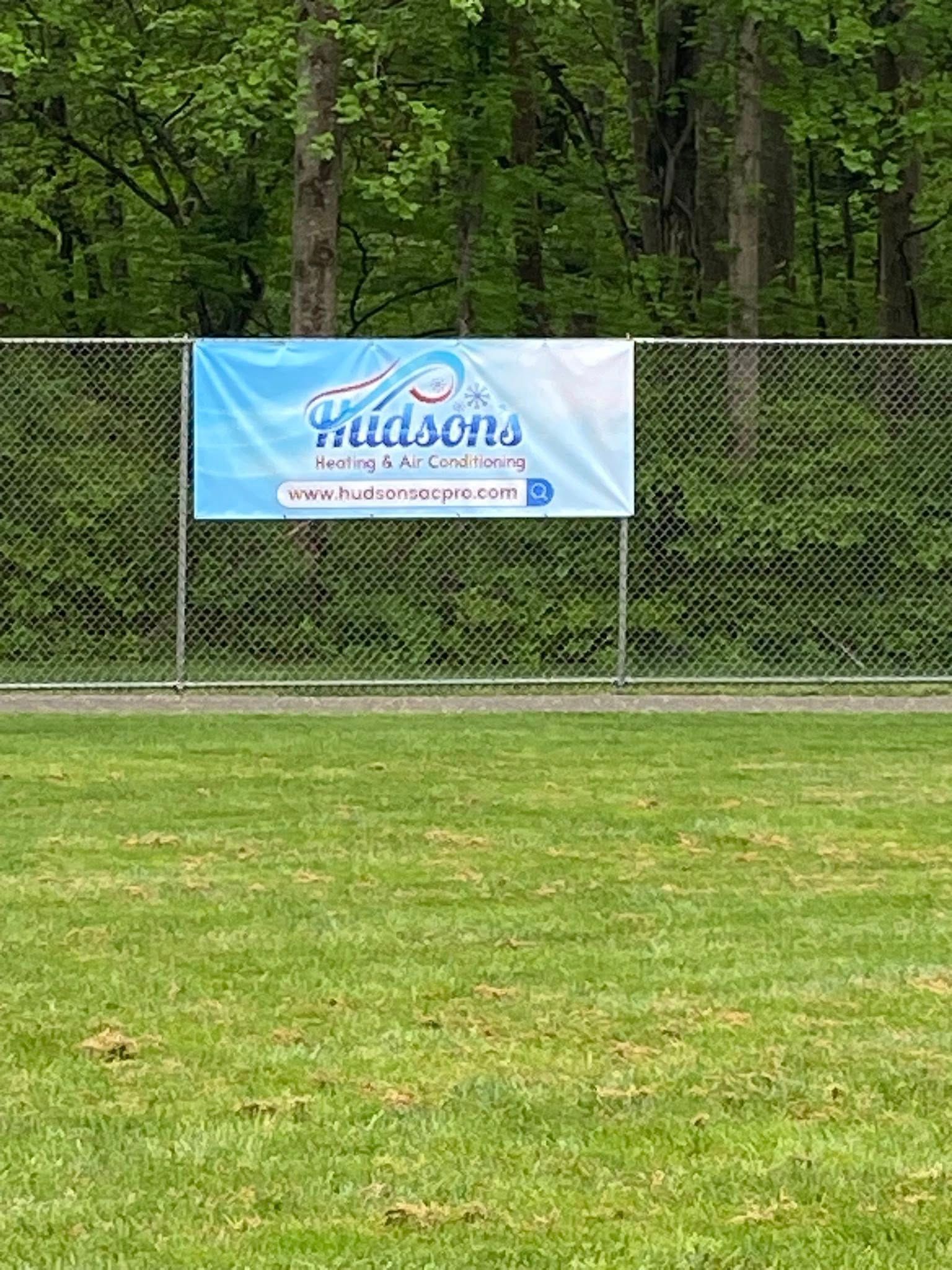 Banner on chain-link fence for Hudson's, likely a business. Blue and white colors; green grass and trees in background.