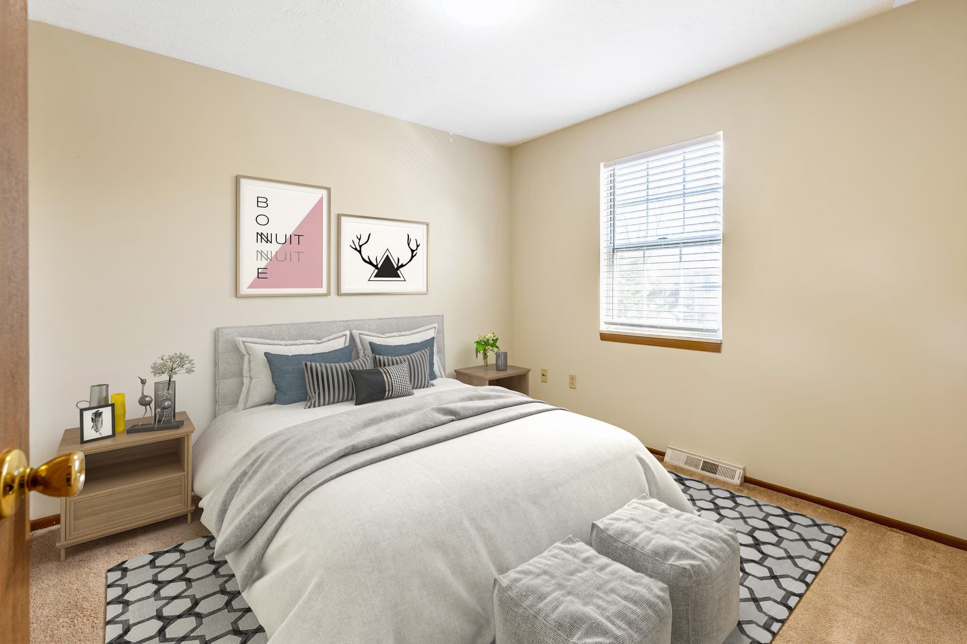 Bedroom with a bed, nightstand, framed art, and a window with blinds. Beige walls and a patterned rug.