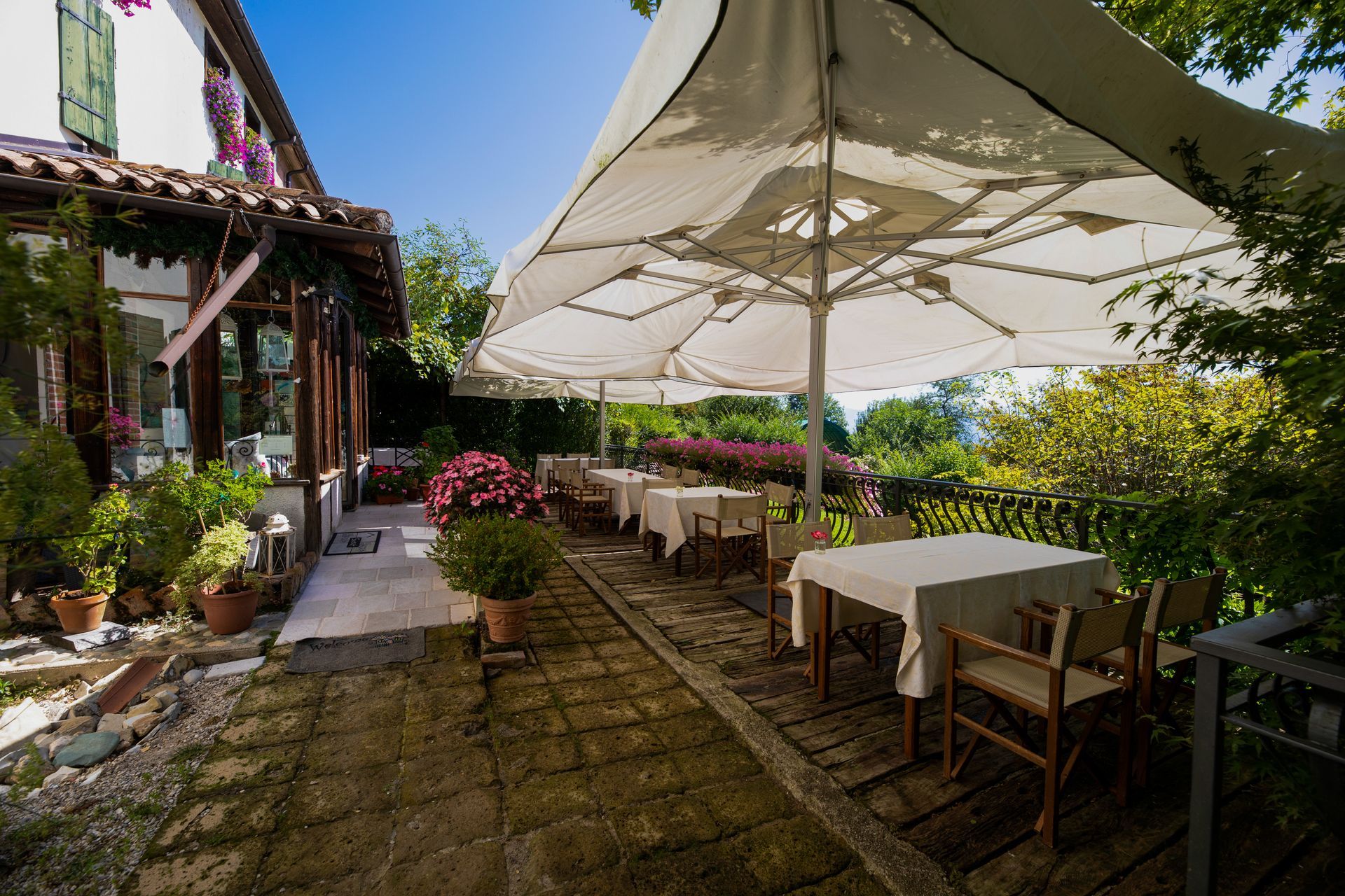 Patio esterno del ristorante con tavoli, sedie e grandi ombrelloni; vegetazione lussureggiante e cielo azzurro.