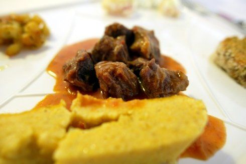 Spezzatino di manzo servito su un piatto bianco con polenta gialla e salsa di pomodoro.