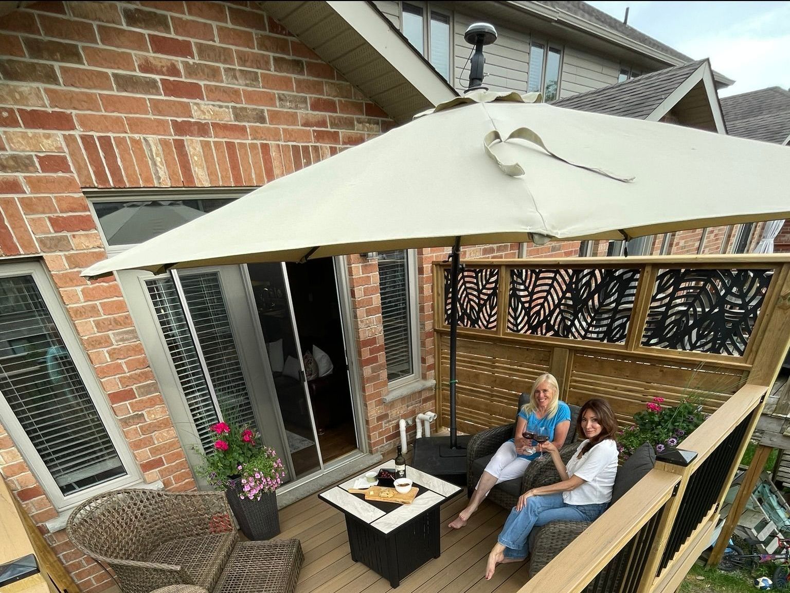 Two women relax on a deck with an umbrella and fire pit, surrounded by a brick building and foliage.