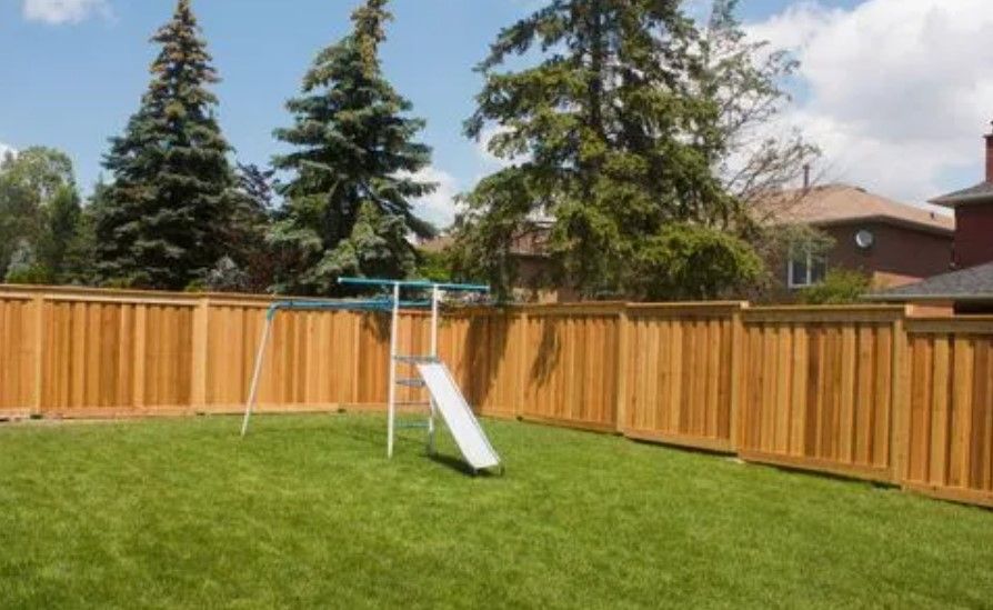 Wooden fence surrounding a backyard with grass, trees, and a small slide.