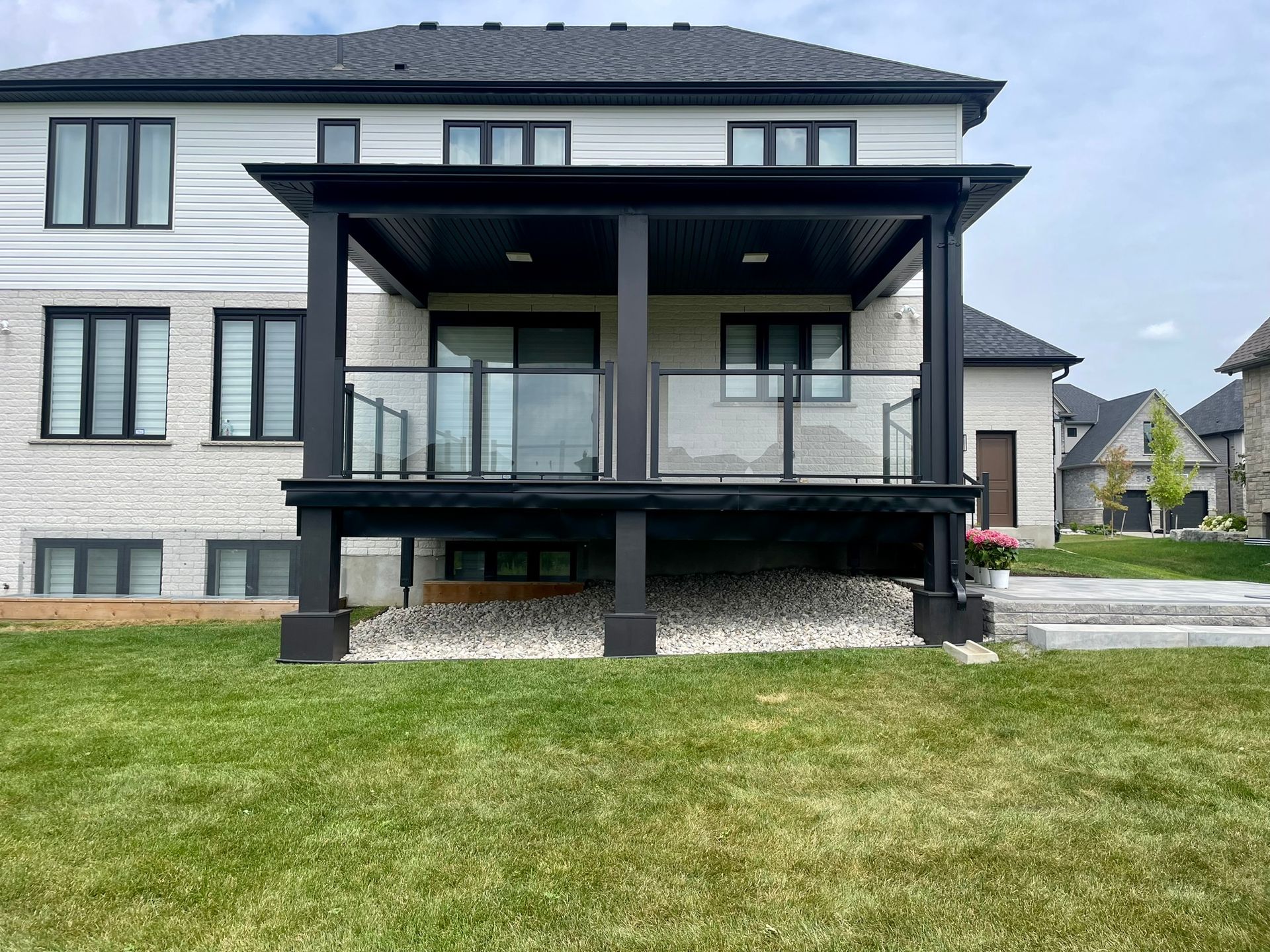 Black deck with glass railings and overhead shelter, attached to a light brick house, on a green lawn.