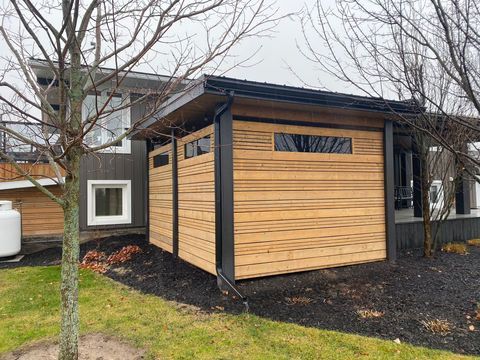 Wooden structure with horizontal slats, attached to a dark-sided house with windows and a gas tank.