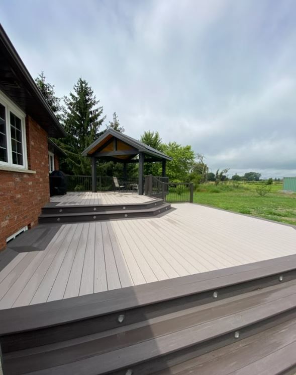 Modern gray deck beside brick house with gazebo, overlooking a green field under cloudy sky
