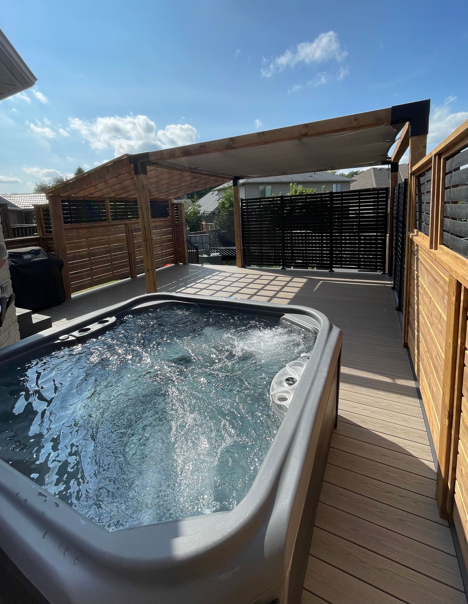 Hot tub bubbling on a wooden patio under a pergola, with a black privacy screen and bright sky beyond.