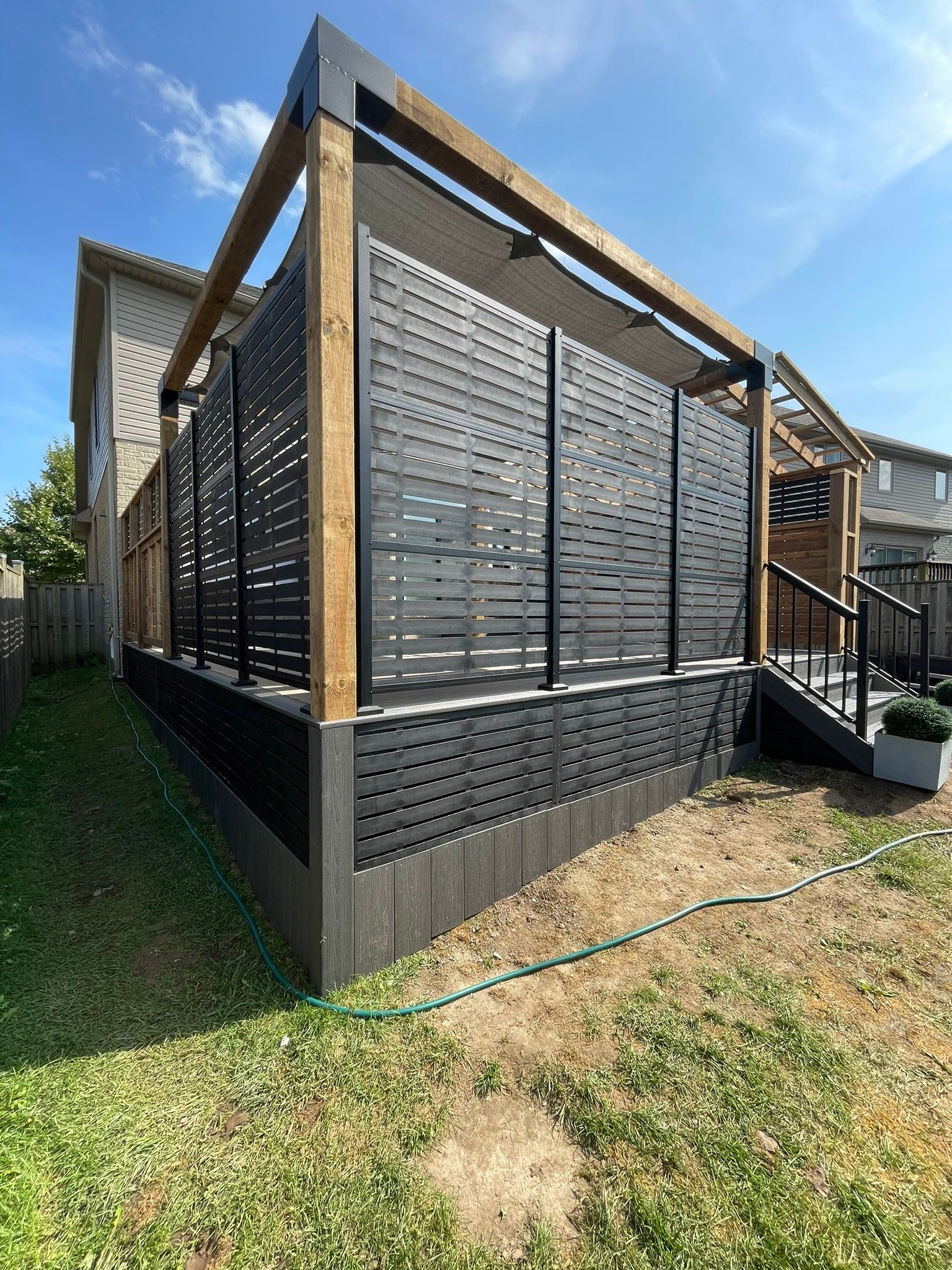 Black and wood outdoor structure with slatted walls, shade, and stairs in a backyard.