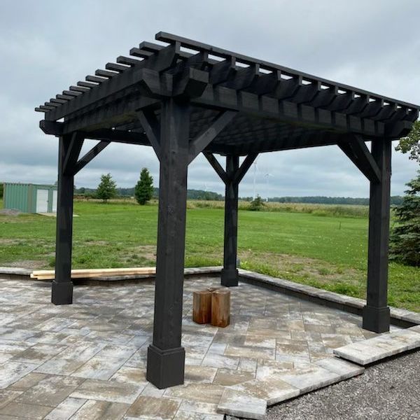 Black wooden pergola on a stone patio, overlooking a field.