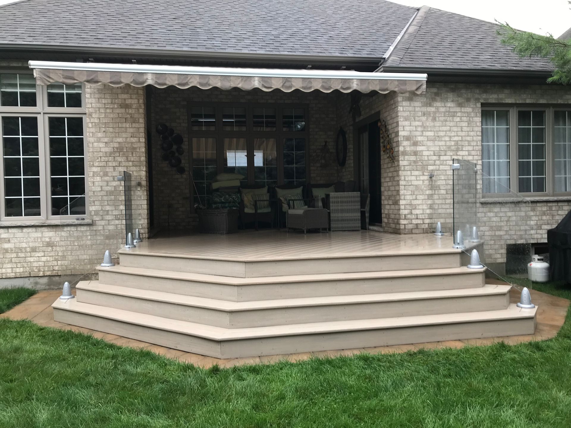 Backyard patio with steps, awning, glass railings, and house in the background.