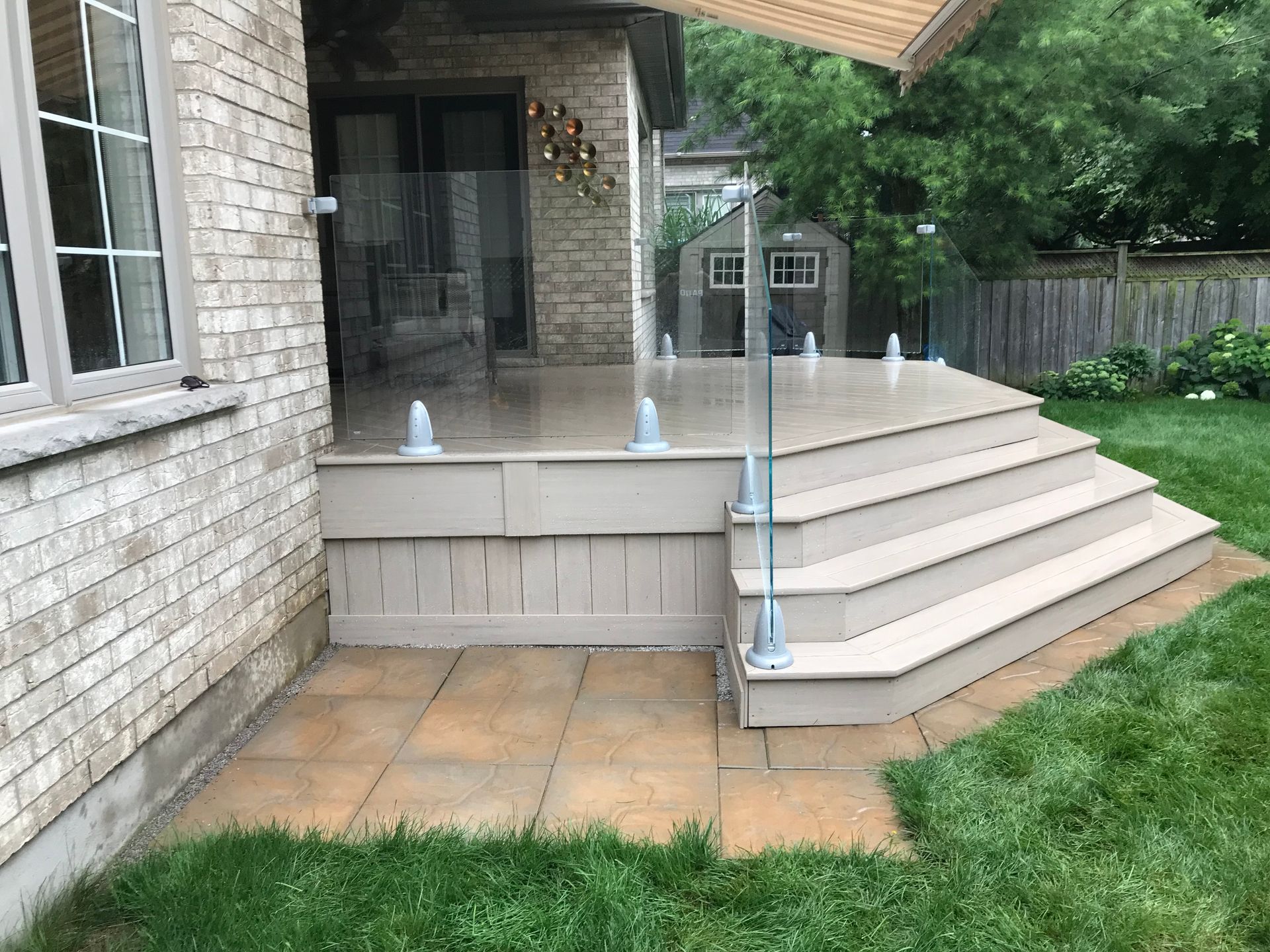 Backyard deck with glass railing and steps leading to patio surrounded by grass.