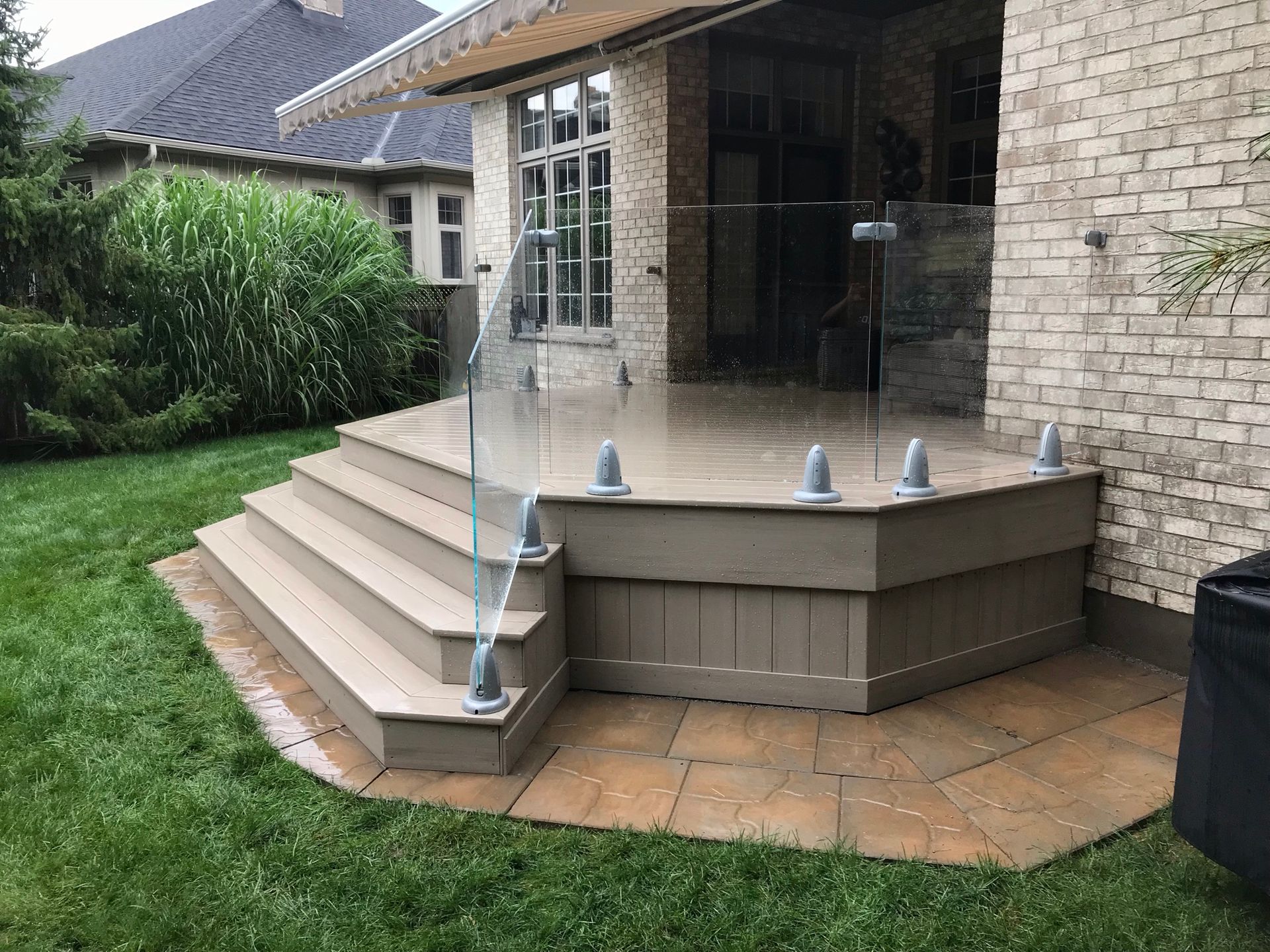 Wooden deck with glass railing, steps, and brick facade. Overlooking a yard with green grass.