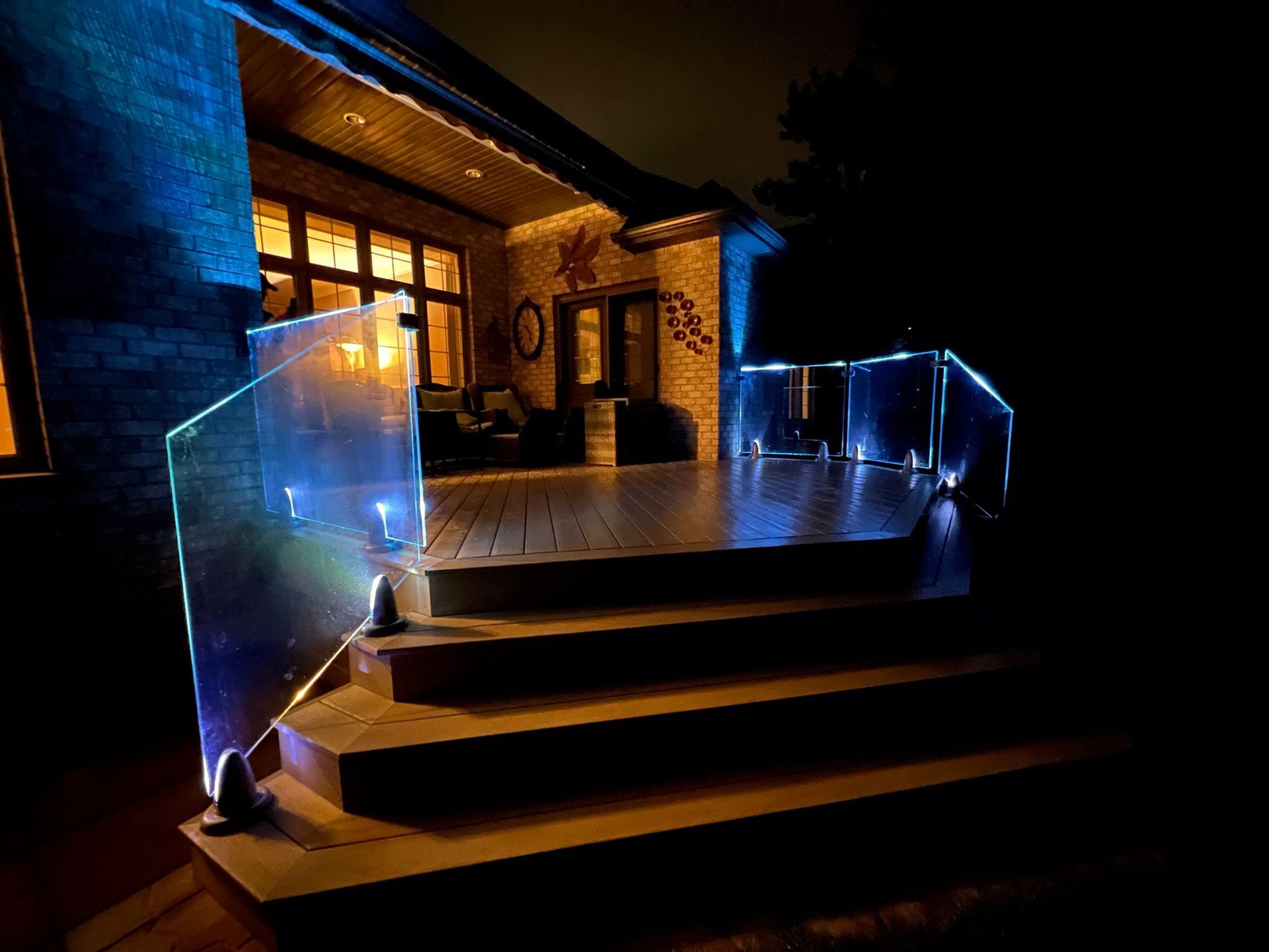 Lit glass deck railing and stairs at night. House with warm interior lights in the background.