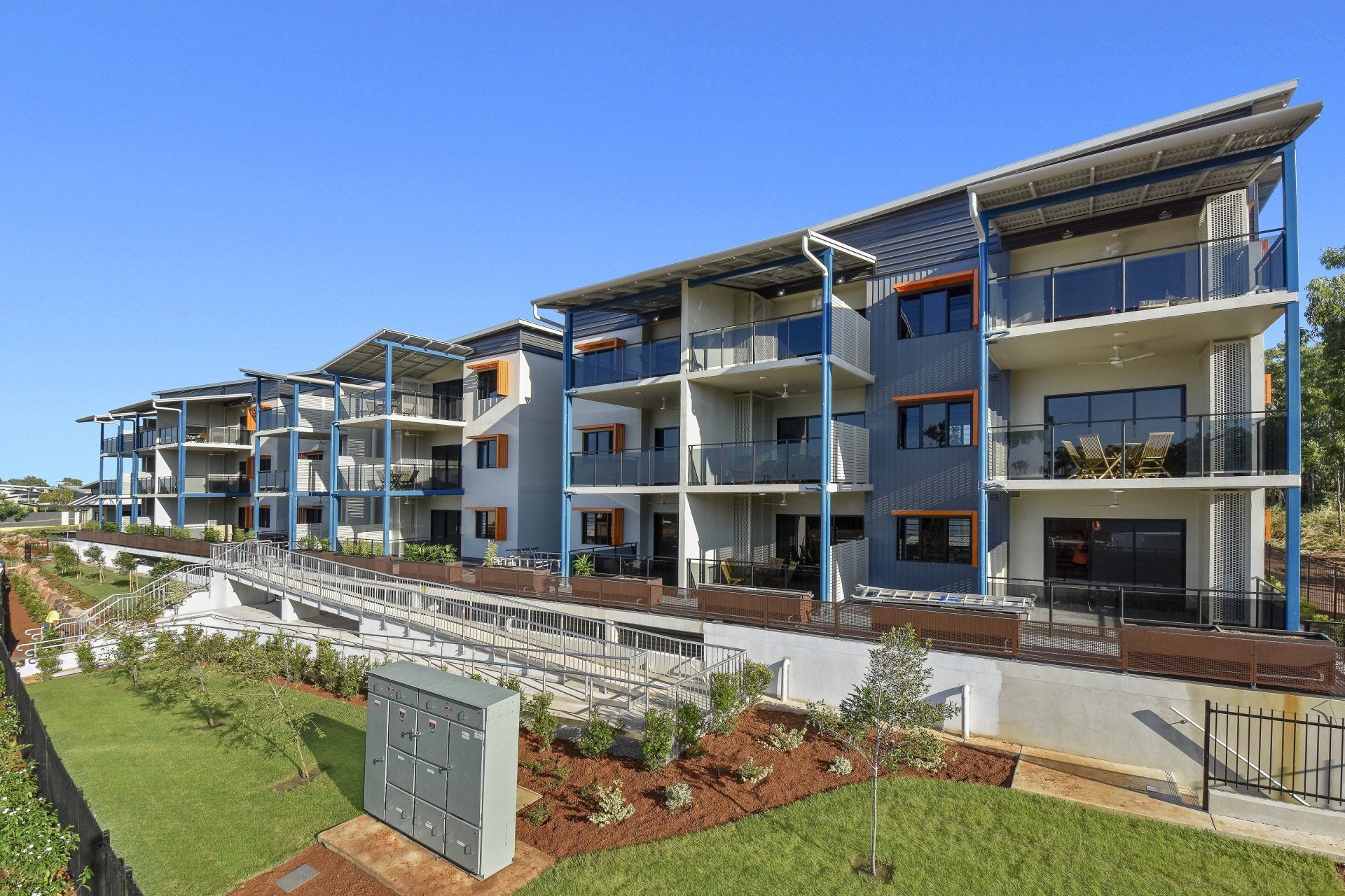 Apartment building with blue and gray facades, balconies, and a grassy area on a sunny day.