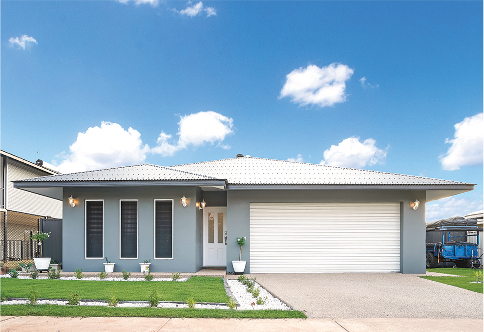 Gray house with white garage door under a blue sky. Front yard with green grass and white stones.
