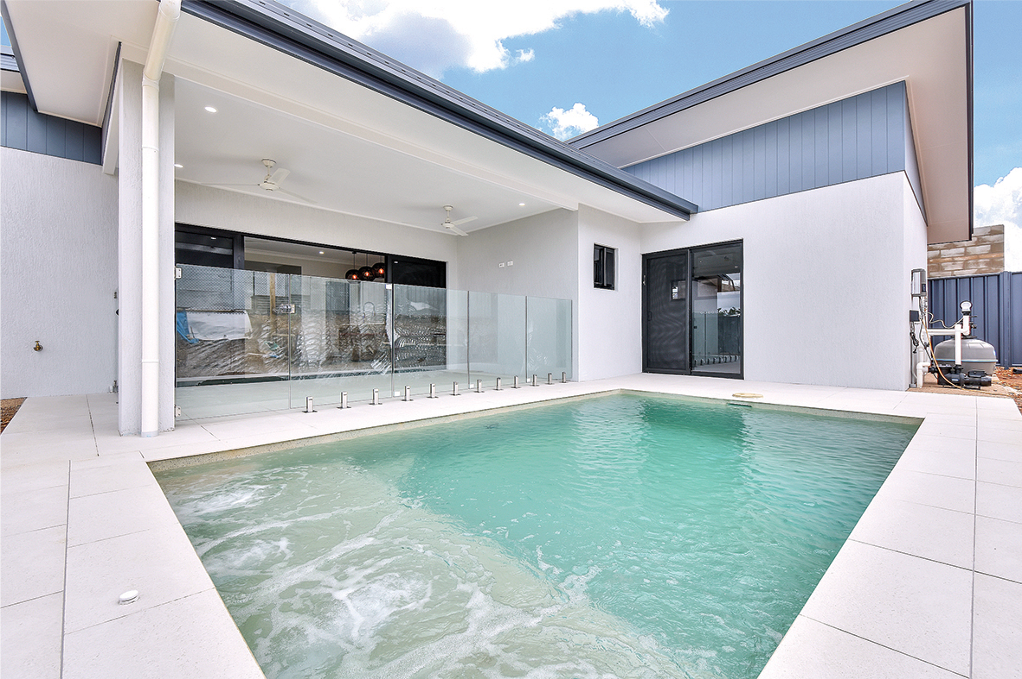 Backyard pool with white patio, glass fence, and house exterior.