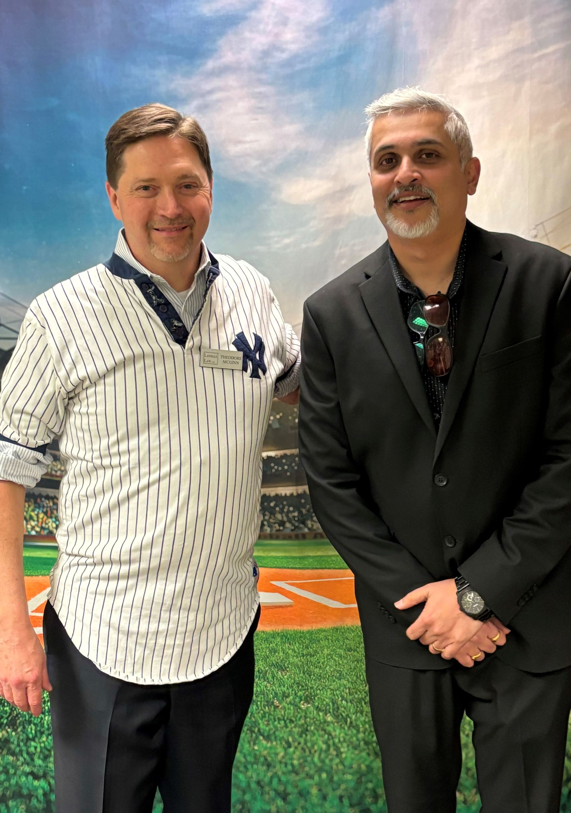 Two men are posing for a picture in front of a baseball field . one of the men is wearing a ny yankees jersey.