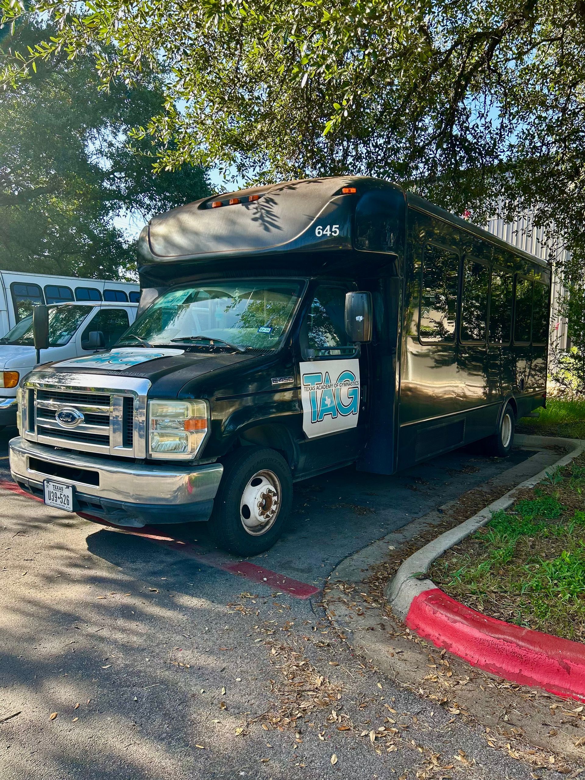 A black shuttle bus with a light blue logo on the door parked in an outdoor lot under trees.