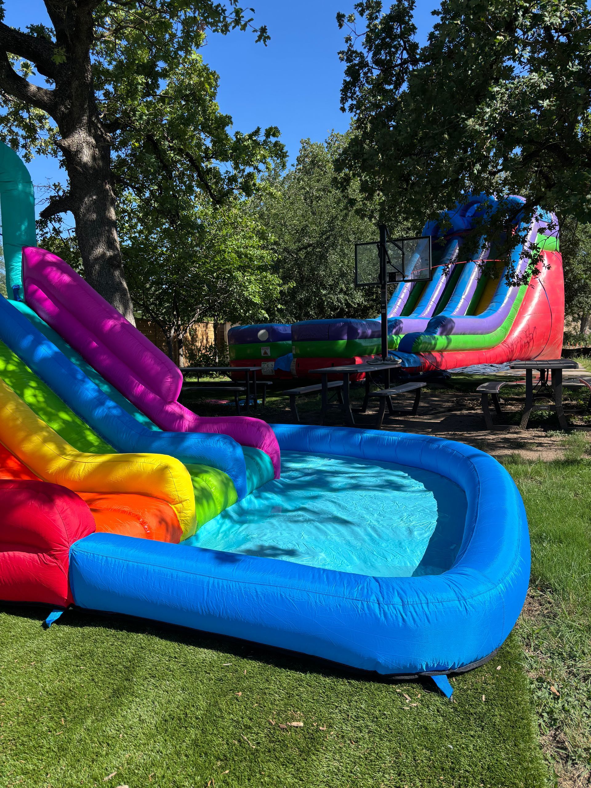 A bright, colorful inflatable water slide with a pool base sits on a grassy lawn in front of a multi-lane bounce house.