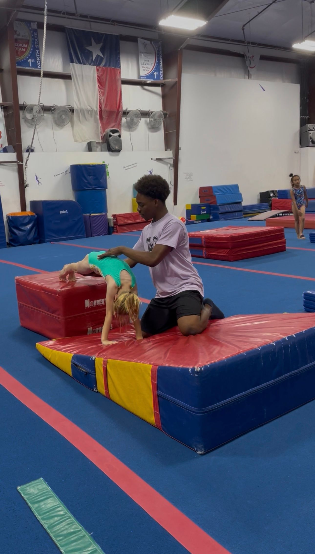 A gymnastics coach spots a student performing a backbend on a red mat inside a gym.