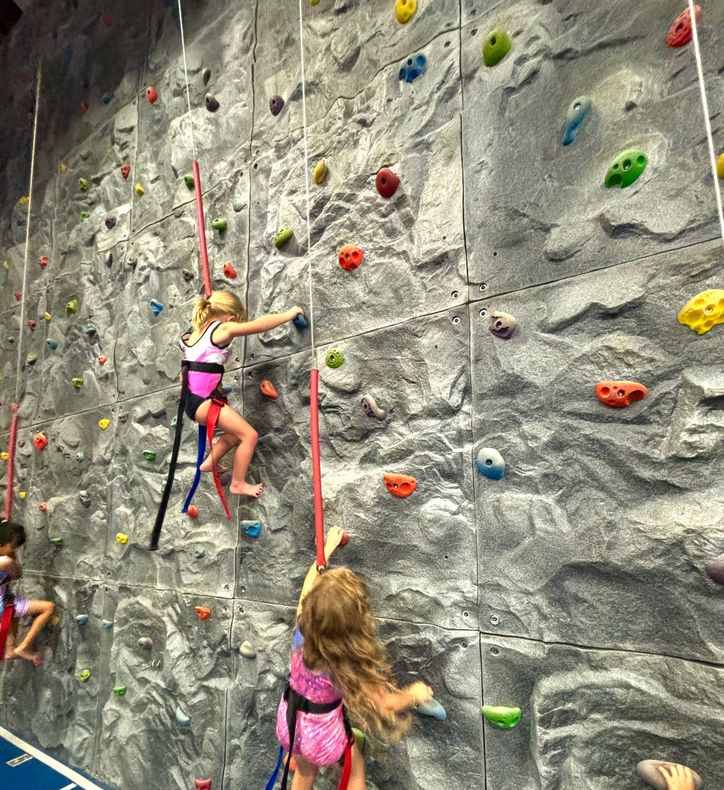 Two children in harnesses climb a rocky indoor climbing wall scattered with various colored grips.