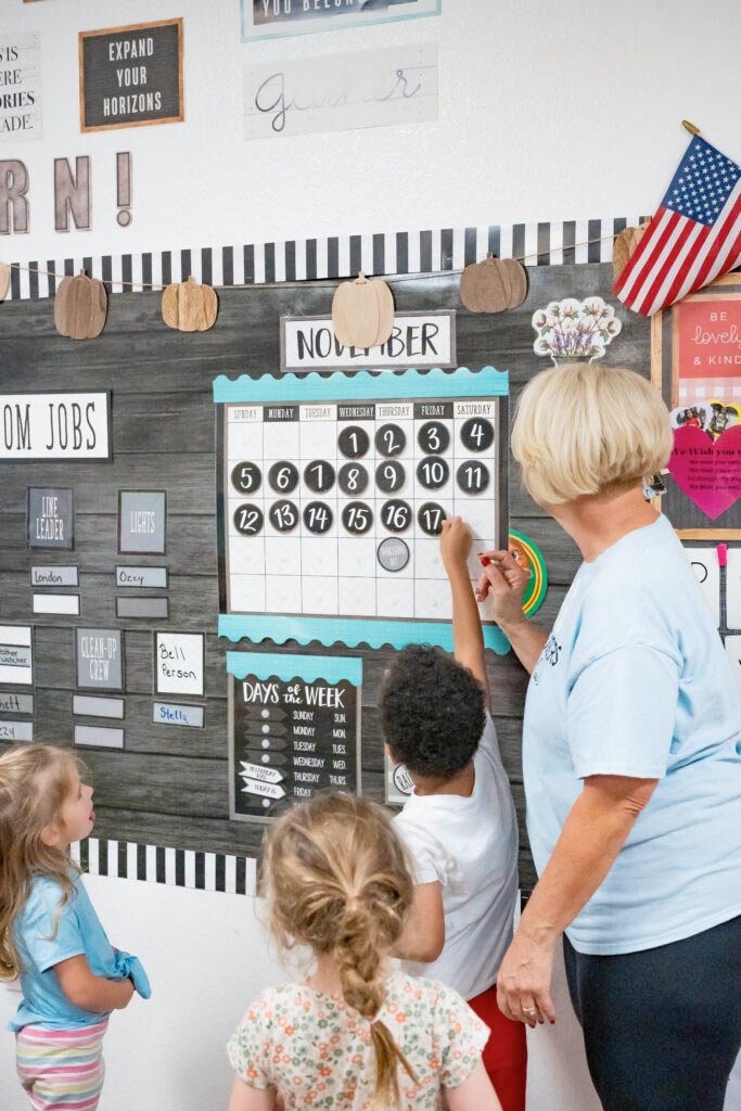 A teacher and three students gather around a classroom calendar on a dark wood-textured wall to look at the date.
