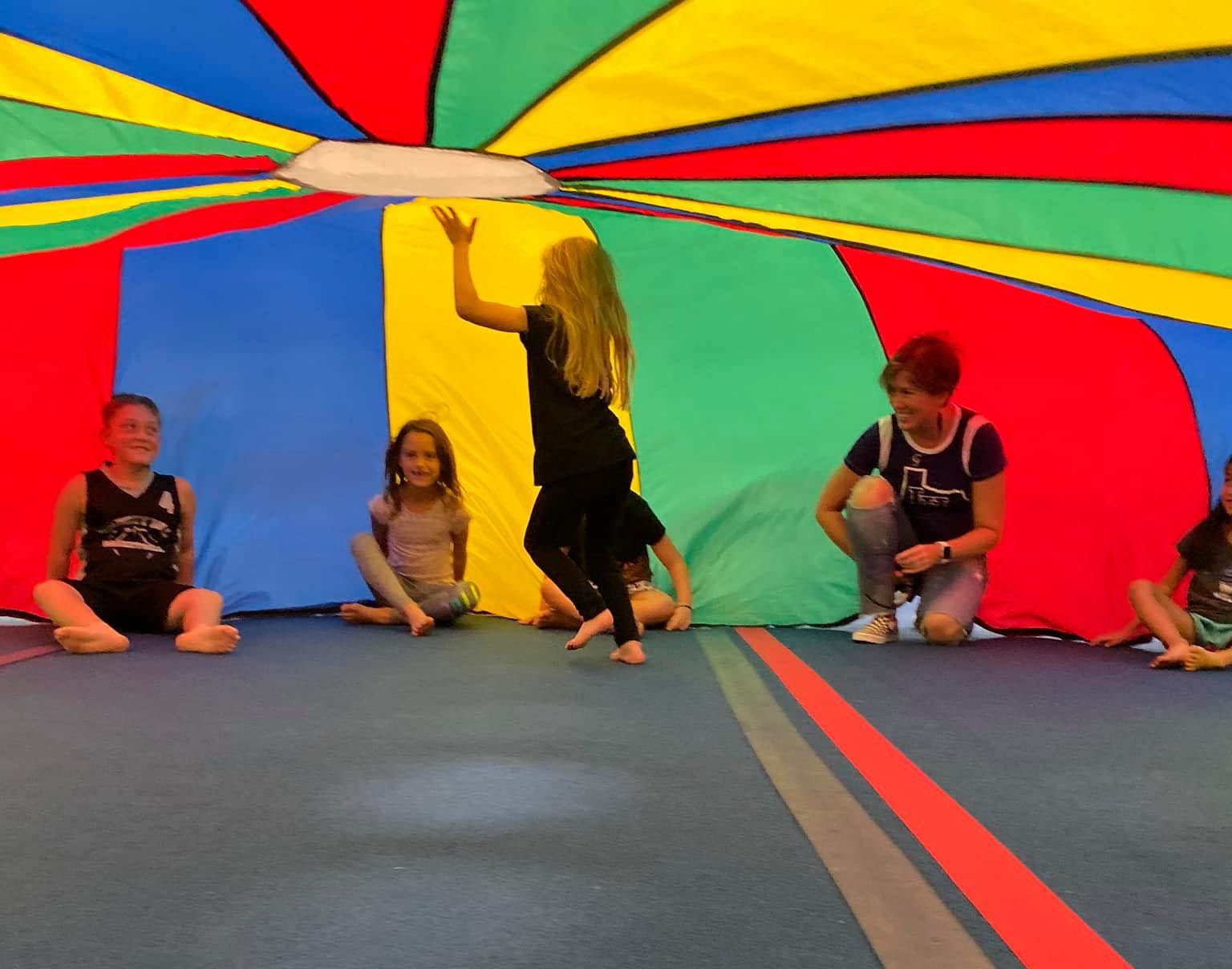 A group of people sit on a blue floor beneath a colorful, circular parachute tent, while one person stands in the center.
