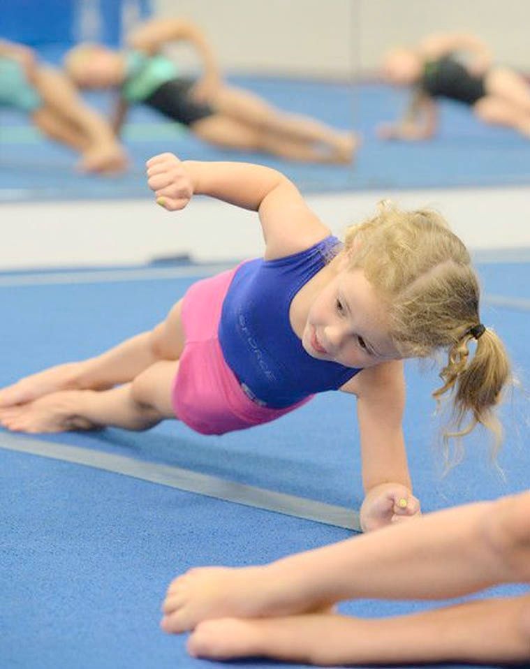 A young gymnast in a blue and pink leotard performs a side plank on a blue mat in a gym.