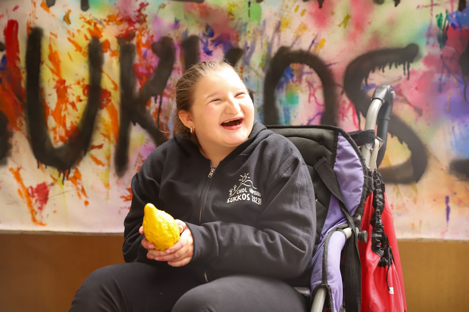 A girl in a wheelchair is holding an esrog and smiling.
