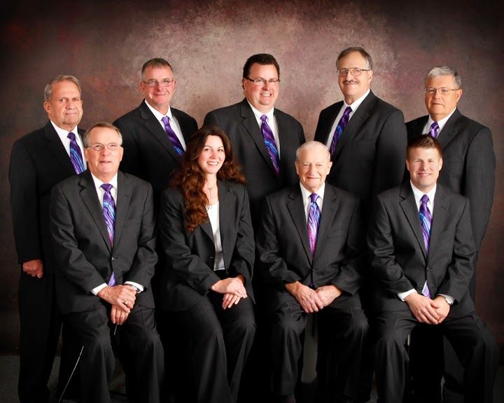 Nine people in black suits pose for a formal group portrait against a studio backdrop.