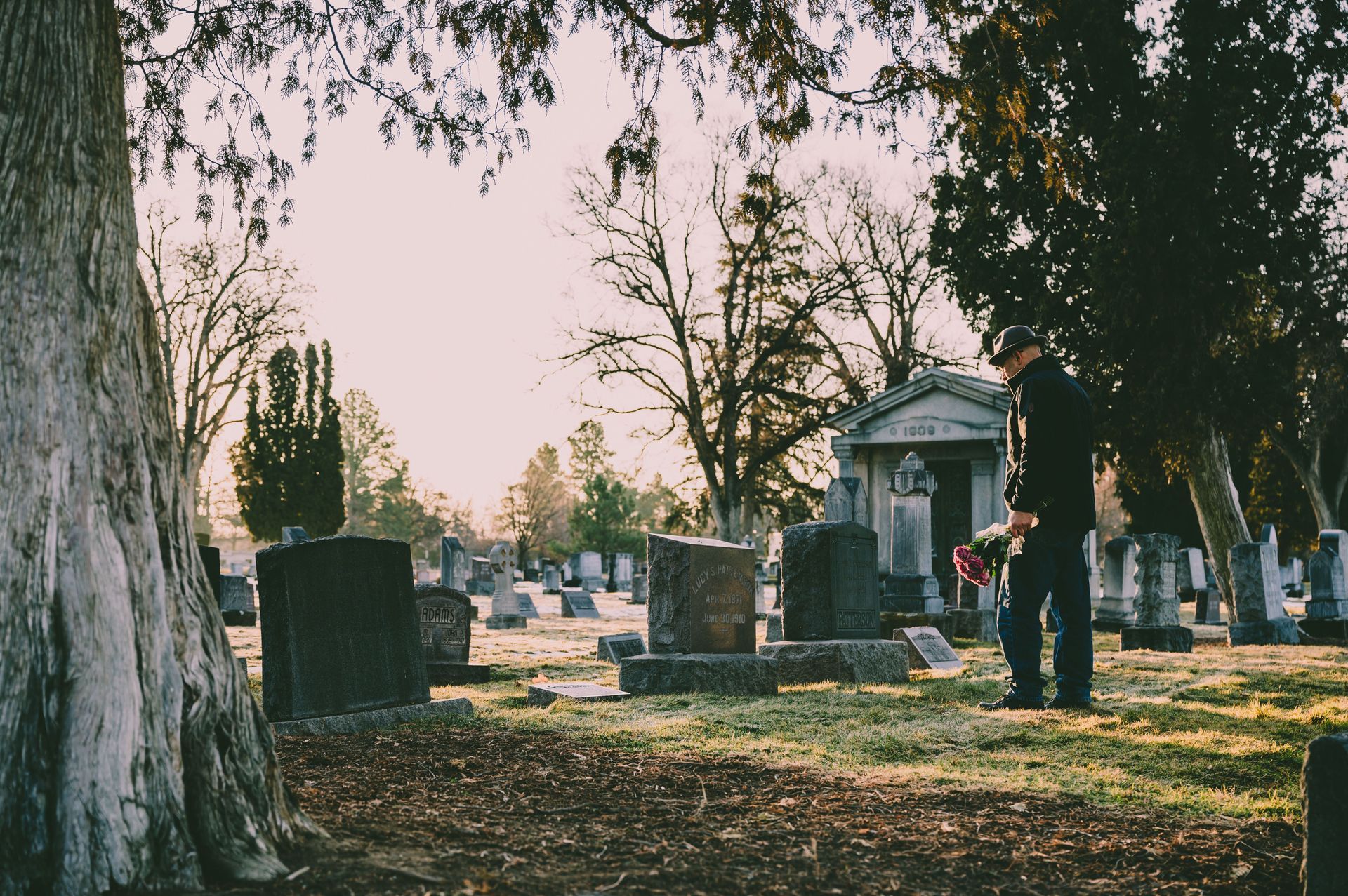 Cemetery scene with a large tree, gravestones, and a person placing flowers near a mausoleum