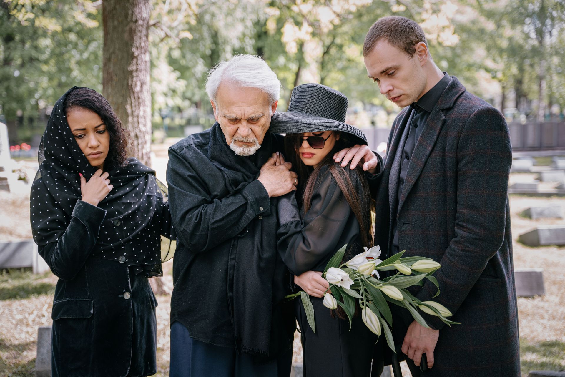 Four people in black mourning clothes stand at a graveside, holding white lilies and embracing in grief.