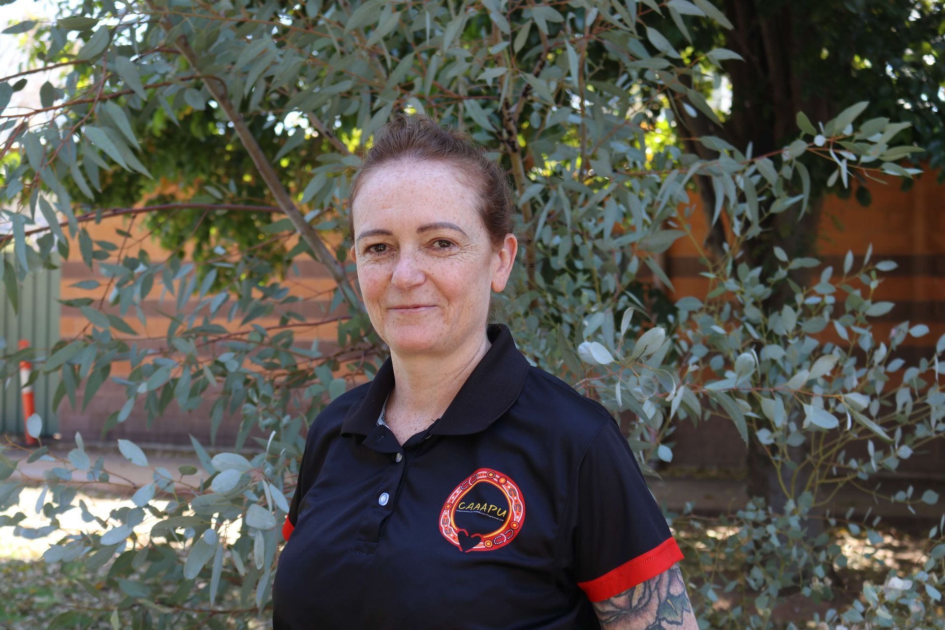 Woman with short brown hair smiles, wearing a light blue button-up shirt, outdoors in front of a tree.