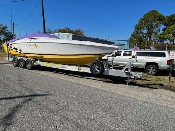 White-and-yellow speedboat on a trailer hitched to a silver pickup in a sunny roadside lot.