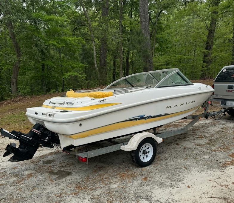 White and yellow boat on a trailer in a wooded gravel lot, with another vehicle partly visible nearby