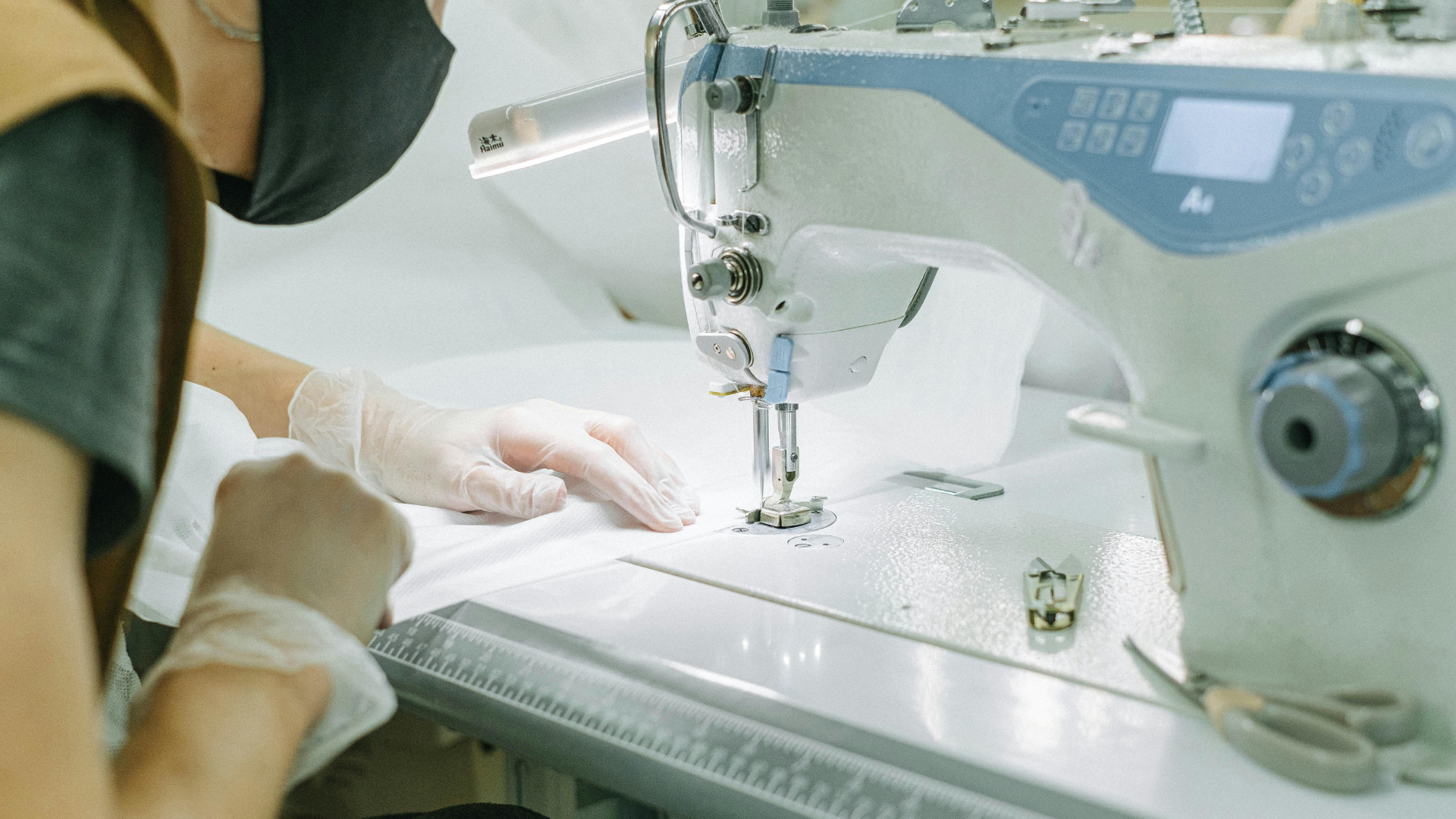 Person wearing mask and gloves sewing white fabric with a sewing machine.