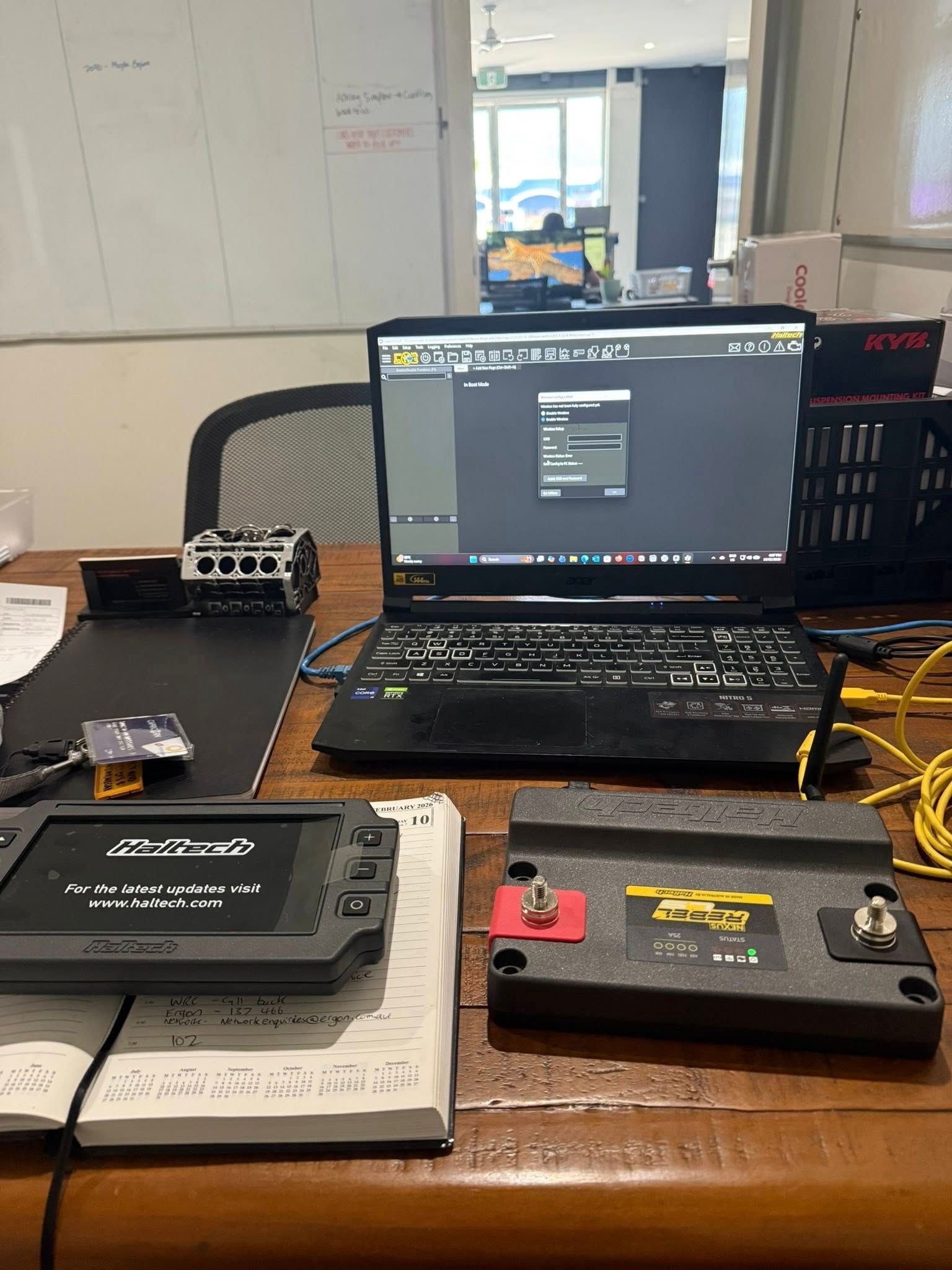 A Laptop Running Software Sits on A Wooden Desk Next to A Battery, a Handheld Device, and Cables in An Office Workspace — Elite Fleet Auto - Electrics & Mechanical in Cannonvale, Qld