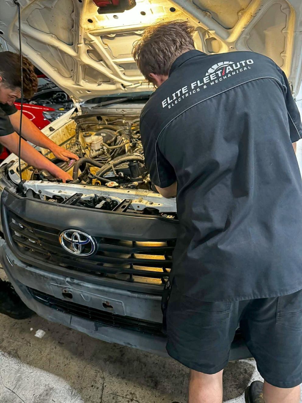 Two Mechanics in Black Uniforms Work on The Open Engine Bay of A Silver Toyota Truck Inside a Garage — Elite Fleet Auto - Electrics & Mechanical in Cannonvale, Qld