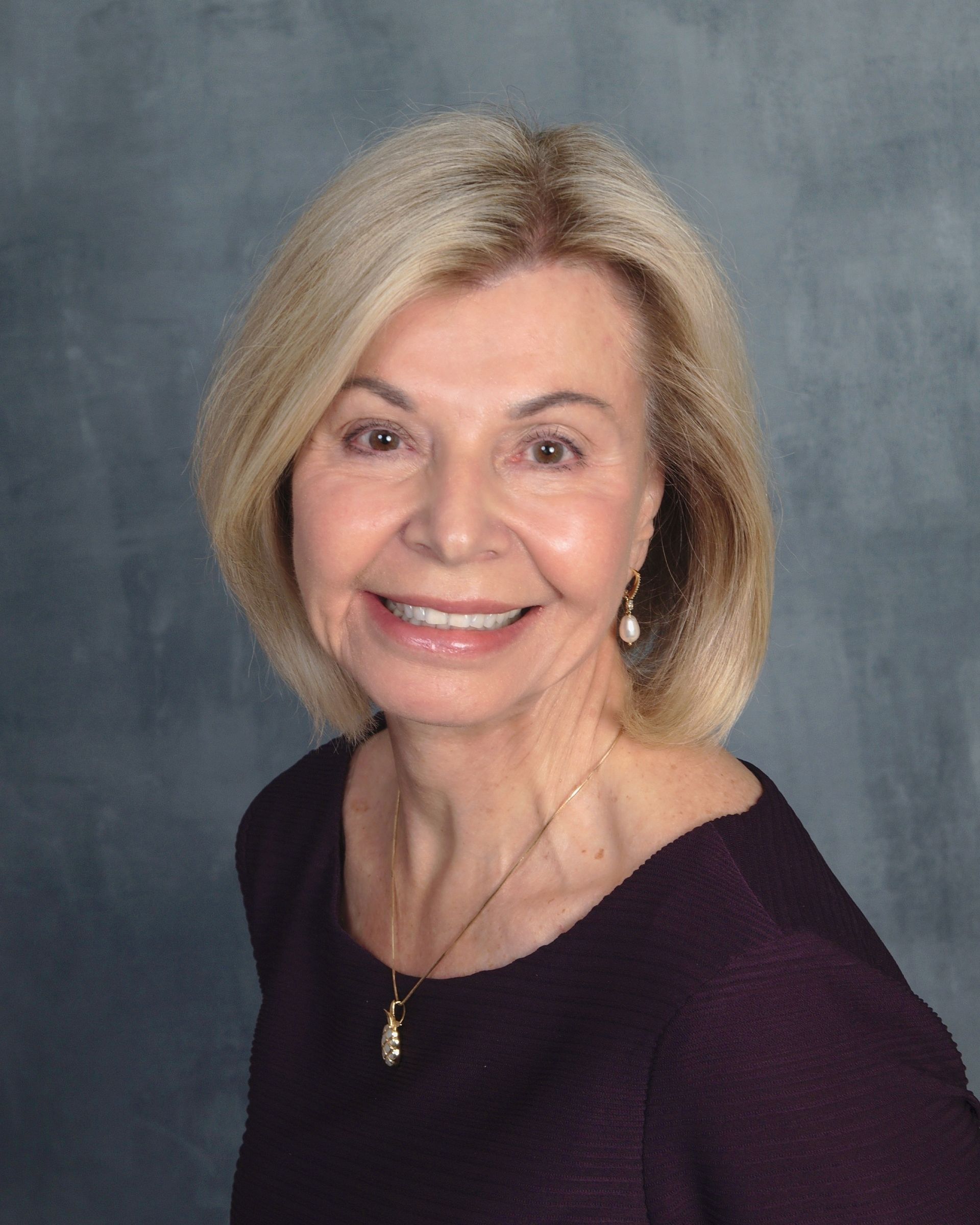 Woman with blonde hair, smiling, wearing a purple top and pearl earrings, set against a blue-gray background.