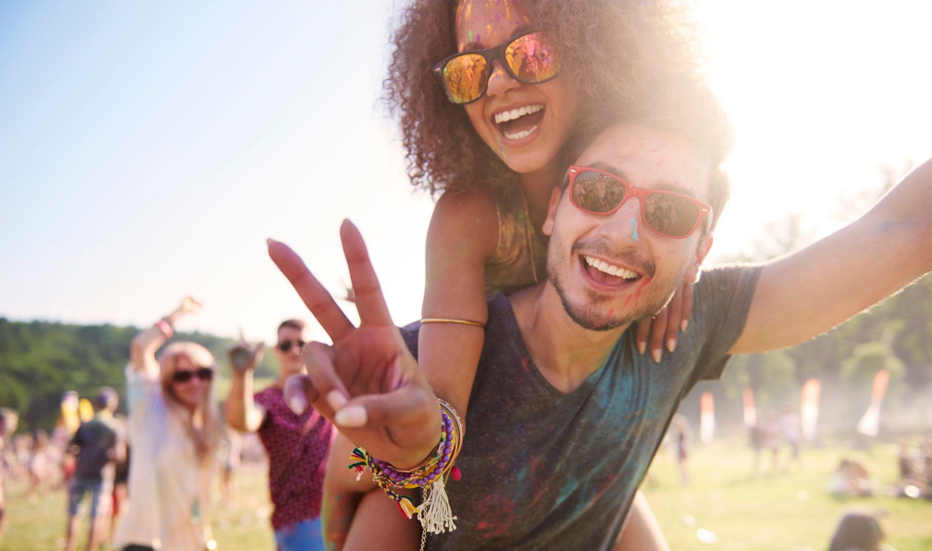 Smiling couple at an outdoor festival; woman on man’s back, sun shining, others in the background.