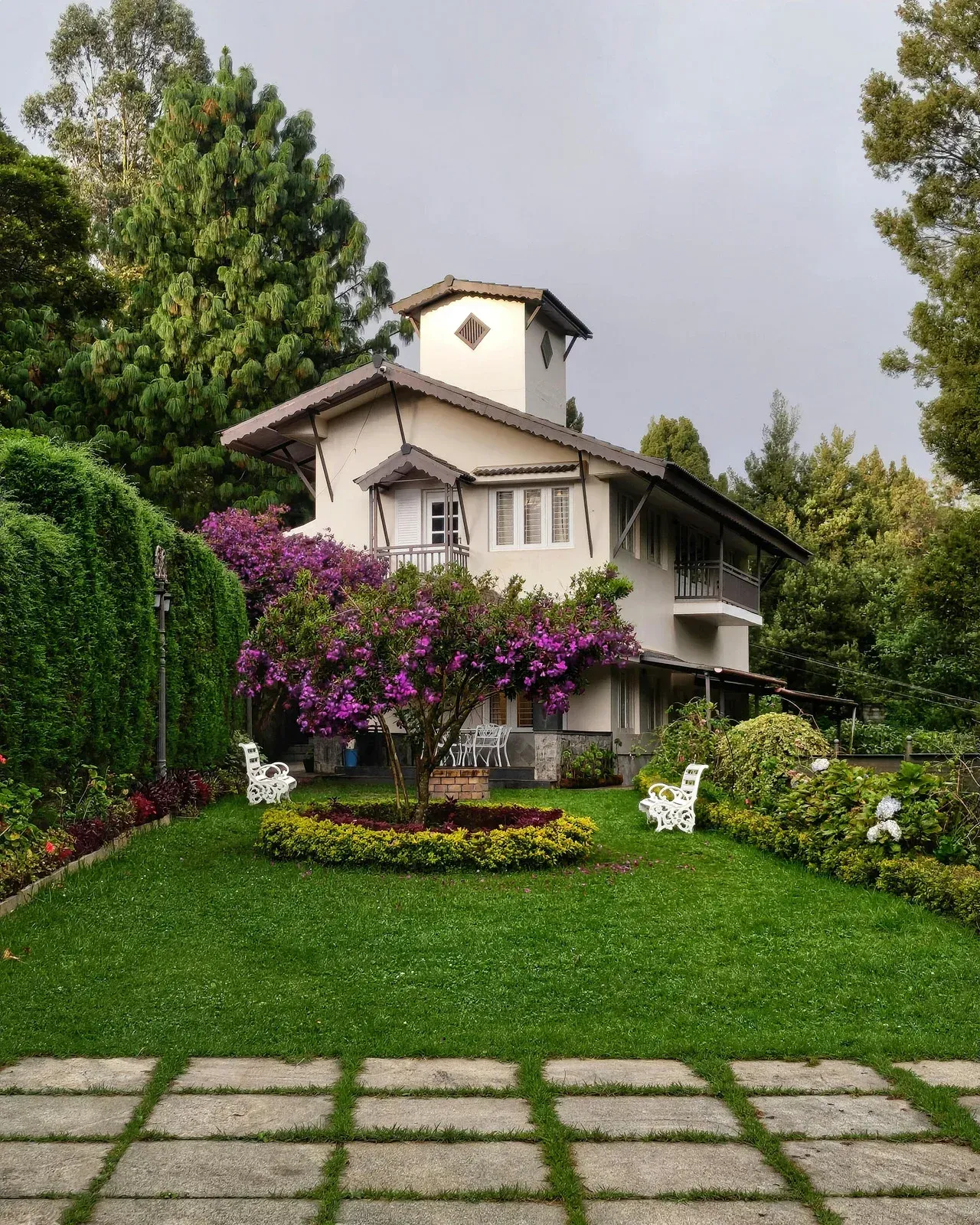 A three-story, light-colored villa stands behind a manicured green lawn with a flowering purple tree and white benches.