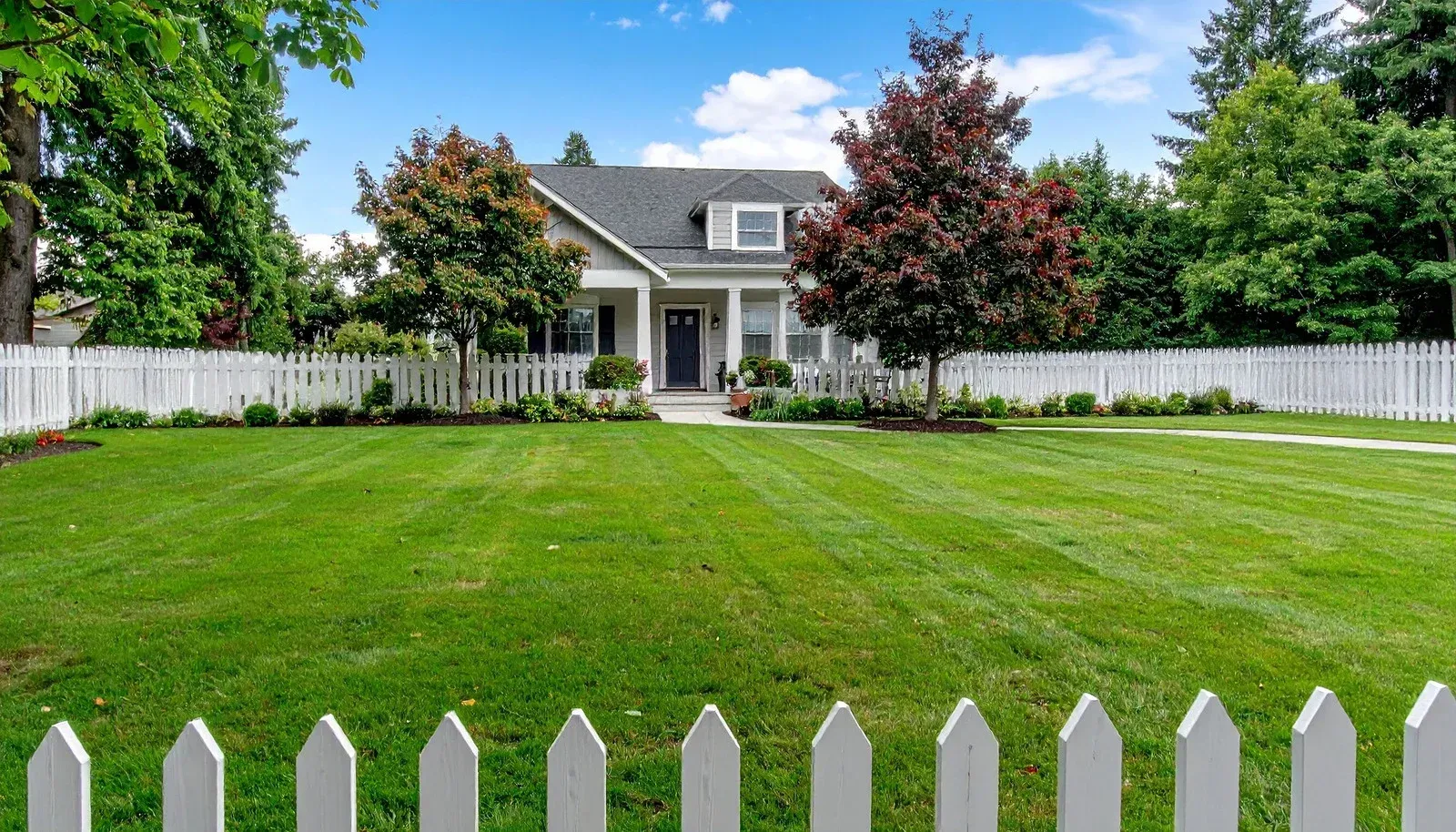 A white picket fence frames a lush green lawn leading to a gray, two-story house surrounded by trees under a blue sky.
