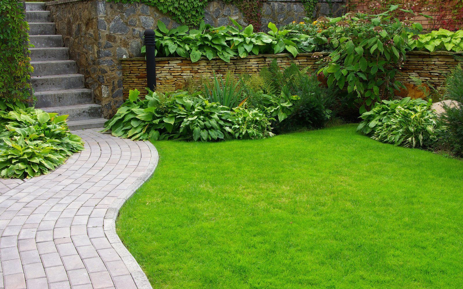 A curved brick walkway leads toward stone steps next to a lush green lawn, low shrubs, and a stone retaining wall.