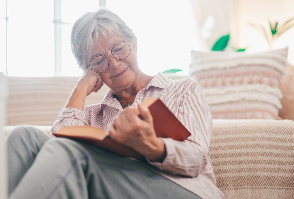 An elderly woman is sitting on a couch reading a book.