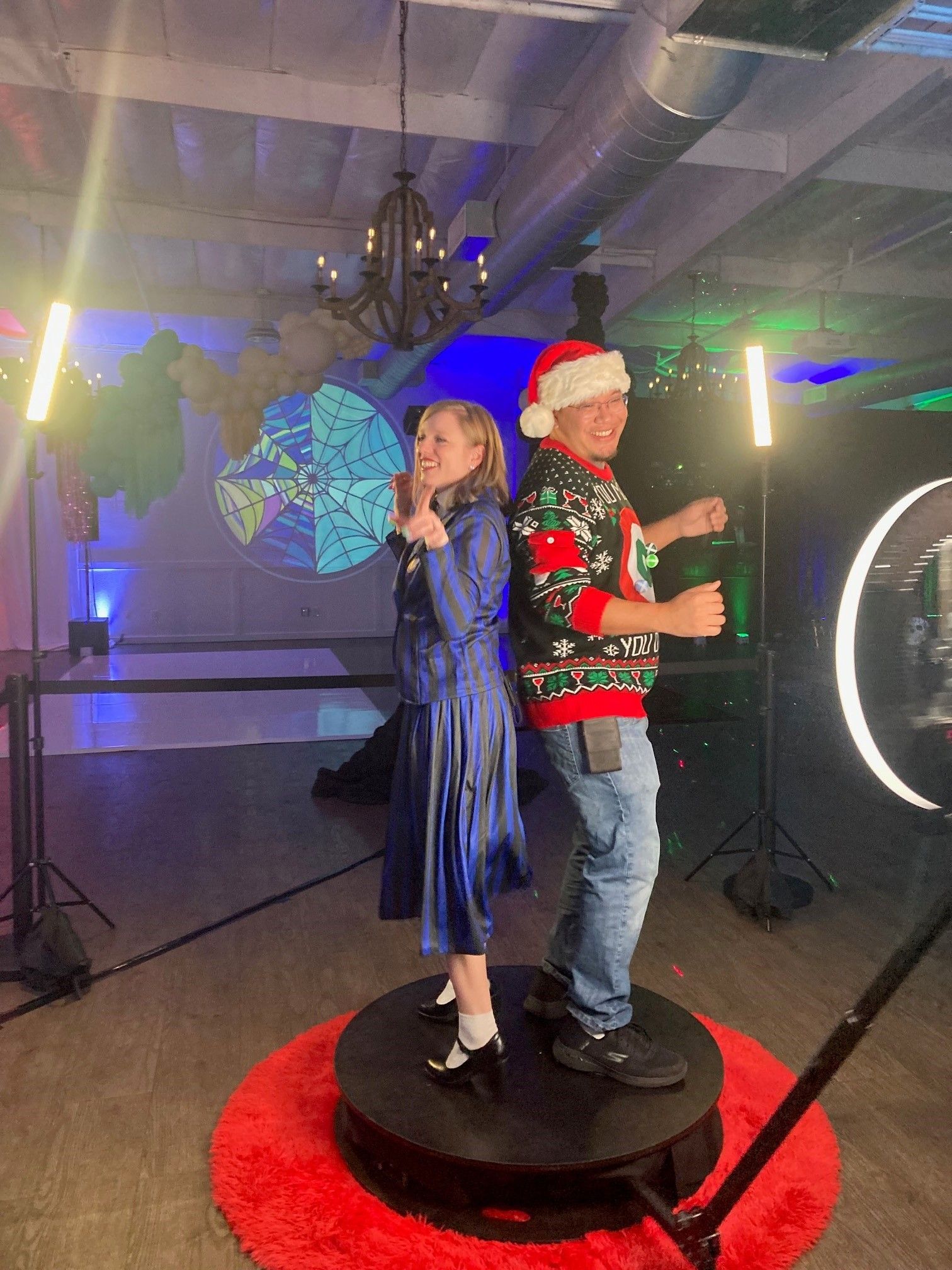 Two performers on a small round stage, one in a blue dress singing and one in a red holiday sweater holding a cup.