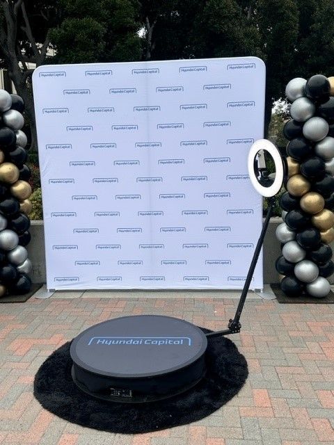 Branded photo backdrop with black-and-white balloon columns and a ring light on a round black platform outdoors