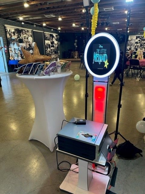 Photo booth kiosk with glowing ring light and sign in a hall, beside a cocktail table display.