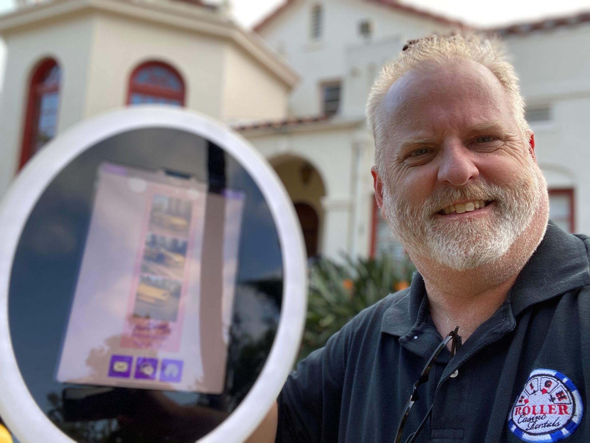 Man smiling beside a sign or phone screen outside a building, holding it up toward the camera