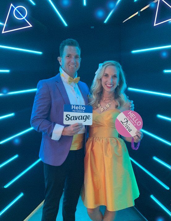 Two people holding signs at a neon blue-lit event backdrop, smiling and posing together.